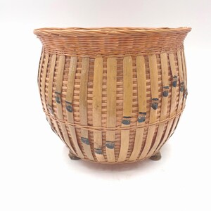 May include: A round, woven basket with a natural brown color. The basket has a flared rim and is supported by three small feet. Vertical bamboo strips are woven into the basket's design, with black painted accents.