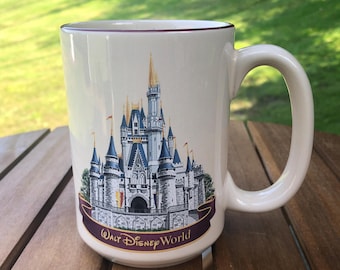 May include: White ceramic mug with a colorful image of Cinderella's Castle from Walt Disney World. The mug has a burgundy rim and handle. The text "Walt Disney World" is printed on the bottom of the mug.