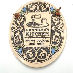 May include: Oval ceramic kitchen sign with the text "GRANDMA'S KITCHEN serves cookies and milk." The sign has a cream-colored background with a decorative black border and blue and green accents. Two small bird-shaped ornaments are attached at the top.