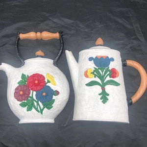 May include: A white ceramic teapot and creamer set with floral designs. The teapot has a black handle and spout, and the creamer has a brown handle. Both pieces feature colorful flowers and leaves.