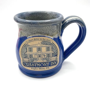 May include: A ceramic mug with a blue handle and a blue and gray rim. The mug features a circular emblem with a drawing of a building and the words "CRESTMONT INN Eagles Mere, PA". The mug also has the text "A short drive to far away."