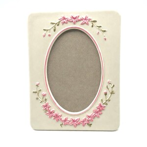 May include: A rectangular off-white picture frame with an oval center opening. The frame features raised relief decorations of pink flowers and green vines arranged in a garland pattern around the top and bottom of the central oval aperture.