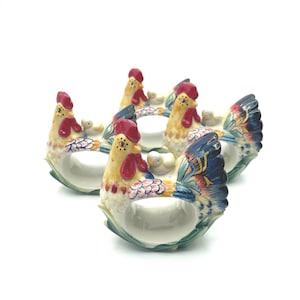 May include: Set of four ceramic rooster napkin rings. Each ring features a colorful rooster design with red combs, yellow bodies, and blue and green tail feathers. The napkin rings are designed to hold a rolled napkin.