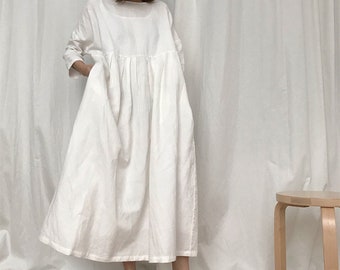 May include: A white linen dress with a loose fit and a gathered waist. The dress has long sleeves and a round neckline. The model is wearing the dress with a pair of light brown ballet flats.