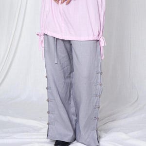 Puede incluir: Una camisa rosa claro de manga larga con cintura con cordón y pantalones grises de pernera ancha. Los pantalones tienen botones decorativos en los laterales. El conjunto está hecho de una tela suave y fluida.