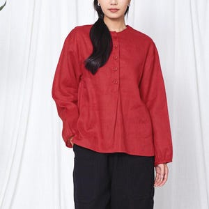 May include: A red long-sleeved shirt with a mandarin collar and button-down front, worn with black trousers. The model is standing in front of a white curtain.