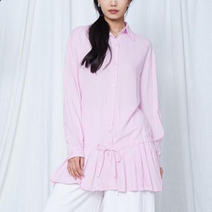 Puede incluir: Una camisa rosa claro con botones, mangas largas y un dobladillo plisado con volantes. La camisa tiene una cintura con cordón y se combina con pantalones blancos de pernera ancha. La modelo lleva la camisa.