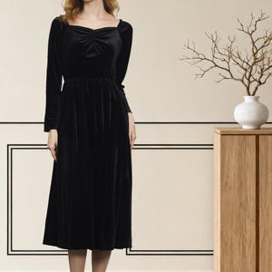 May include: A long-sleeved black velvet dress with a square neckline and a gathered waist. The dress falls to mid-calf and is paired with black heels. The background features a neutral wall with black trim and a wooden cabinet with a white vase.
