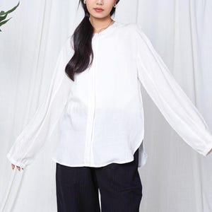Può includere: Una camicia bianca a maniche lunghe con colletto alla coreana e abbottonatura. Indossata con pantaloni neri. La modella è in piedi davanti a una tenda bianca, con foglie verdi sullo sfondo.