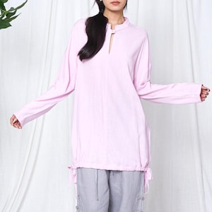 May include: A light pink long-sleeve tunic with a keyhole neckline and drawstring detail at the waist. The tunic is paired with gray pants. The outfit is made of a soft, flowing fabric.