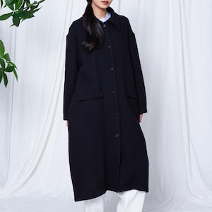May include: A long, dark navy coat with a button-down front and two front pockets. The coat is worn over a light blue collared shirt and white pants. The model is wearing black shoes. The background is a white curtain.
