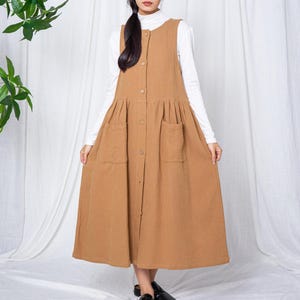 Könnte beinhalten: Ein beigefarbenes Knopfkleid mit zwei Vordertaschen und einem ausgestellten Rock. Das Kleid wird über einem weißen Rollkragenpullover mit langen Ärmeln getragen. Das Kleid ist knielang und hat ein schlichtes, klassisches Design.