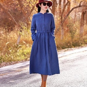 Peut inclure: Une femme portant une robe en lin bleu avec un chapeau rouge et des lunettes de soleil se tient sur une route pavée avec des arbres en arrière-plan.