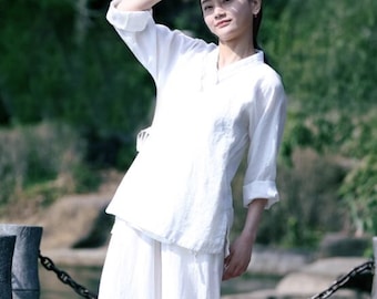 May include: A woman wearing a white linen pantsuit. The pants are wide-legged and the top has a mandarin collar. She is standing on a wooden walkway with a chain link fence and a body of water in the background.