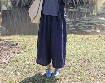 May include: A woman wearing a blue and white striped long-sleeve shirt, navy blue culottes, and blue shoes. She is carrying a beige tote bag and a black vest. The background features a pond and lush greenery.