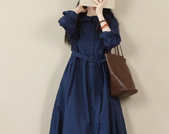 May include: A navy blue long-sleeved dress with a white collar and a belt. The dress has a button-up front and a slightly flared skirt. The model is wearing white sneakers and a brown tote bag.