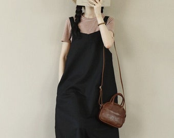 May include: A woman wearing a black linen overall dress with a brown leather crossbody bag and a straw hat. She is holding a book in front of her face.