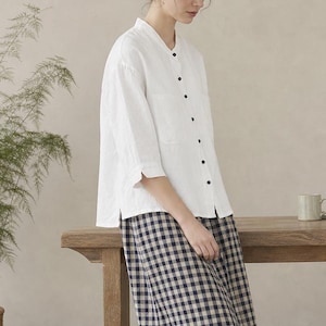 May include: A white button-down shirt with a mandarin collar and three-quarter length sleeves paired with a navy blue and white gingham midi skirt. The outfit is completed with brown leather sandals featuring crisscross straps over the feet.