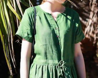 May include: A woman wearing a green linen dress with a button-up front and a tie at the waist. The dress has short sleeves and a v-neck.