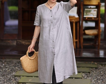 May include: A gray linen shirt dress with a button-up front and a side slit. The dress has short sleeves and a relaxed fit. The model is wearing black pants and holding a woven basket bag.
