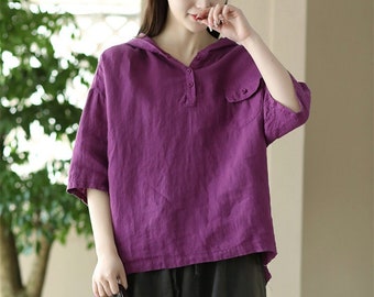 May include: A woman's outfit featuring a purple hooded top with short sleeves and a pocket, paired with dark gray, loose-fitting pants. A woven straw tote bag with leather handles completes the ensemble. The outfit is casual and comfortable.