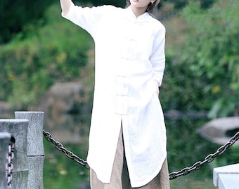 May include: A woman wearing a white linen tunic top with a mandarin collar and brown linen pants. She is wearing a beige cap and standing on a stone bridge over a pond.