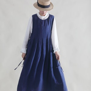 Puede incluir: Un vestido de lino azul marino sin mangas con una falda plisada. El vestido tiene tirantes ajustables que se atan a los lados. La modelo lleva una blusa blanca debajo del vestido y un sombrero de paja con una cinta negra.