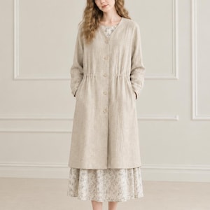 May include: A beige long-sleeved coat with a V-neckline and a button-down front, featuring a cinched waist with an internal drawstring. Underneath, a white midi dress with a delicate grey floral pattern is visible, paired with beige loafers.