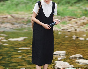 May include: A woman wearing a black linen dress with a white ruffle collar stands in a shallow stream. She is holding a bottle in her right hand. A wicker basket with a watermelon and other items is in the water behind her.