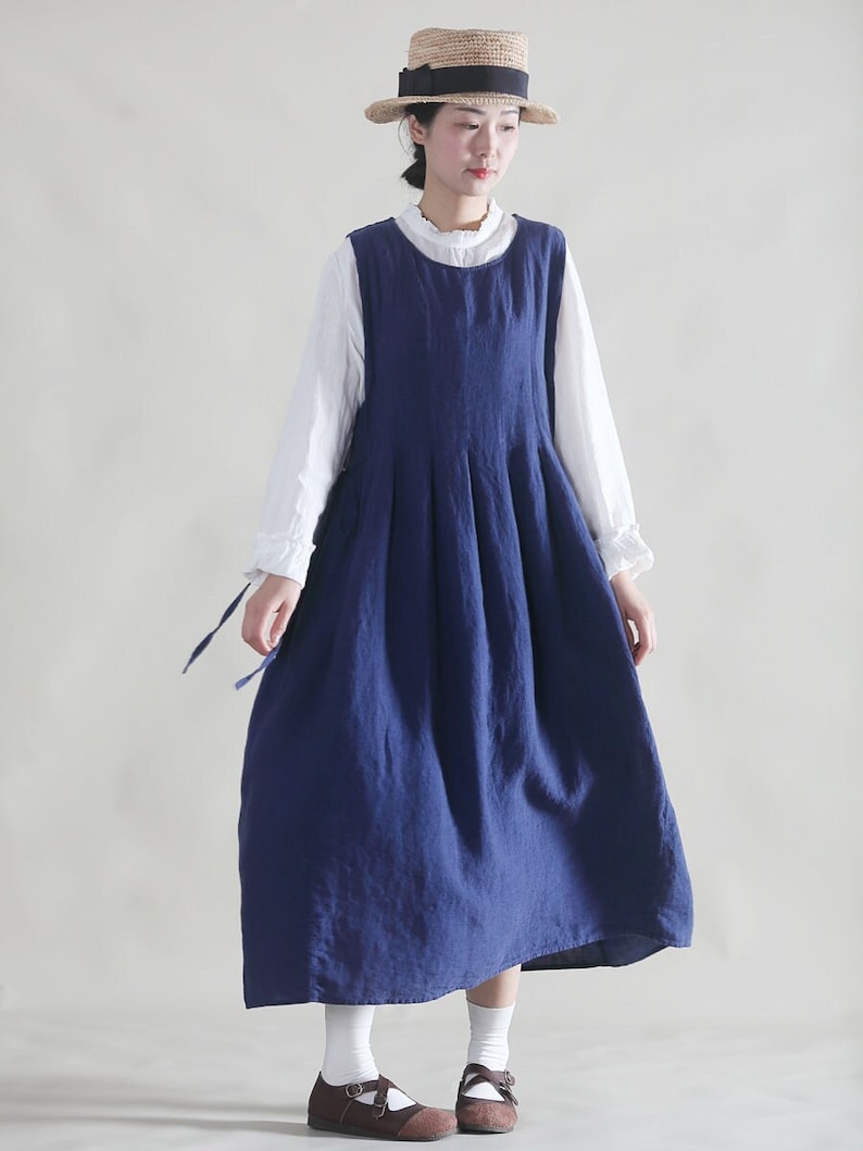 Puede incluir: Un vestido azul de lino sin mangas con una falda plisada y una blusa blanca debajo. La modelo lleva un sombrero de paja con una cinta negra.