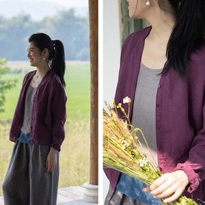 May include: A woman wearing a purple linen cardigan with a button-up front. The cardigan has long sleeves and a relaxed fit. The woman is standing in a field with a bouquet of flowers in her hand.