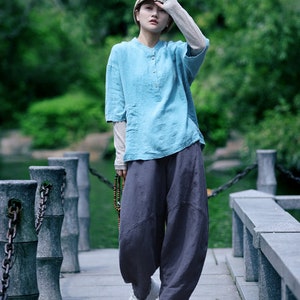May include: A person wearing a light blue linen shirt with a mandarin collar and dark gray, loose-fitting pants. The outfit is paired with white platform sneakers and a beige cap. The person is standing on a stone walkway.