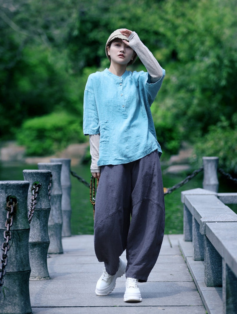 May include: A person wearing a light blue linen shirt with a mandarin collar and dark gray, loose-fitting pants. The outfit is paired with white platform sneakers and a beige cap. The person is standing on a stone walkway.