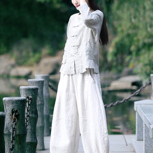 May include: A person wearing a white linen outfit, including a long-sleeved top and wide-leg pants. The outfit is paired with a matching head covering and white sneakers. The person is standing on a stone walkway near a body of water.