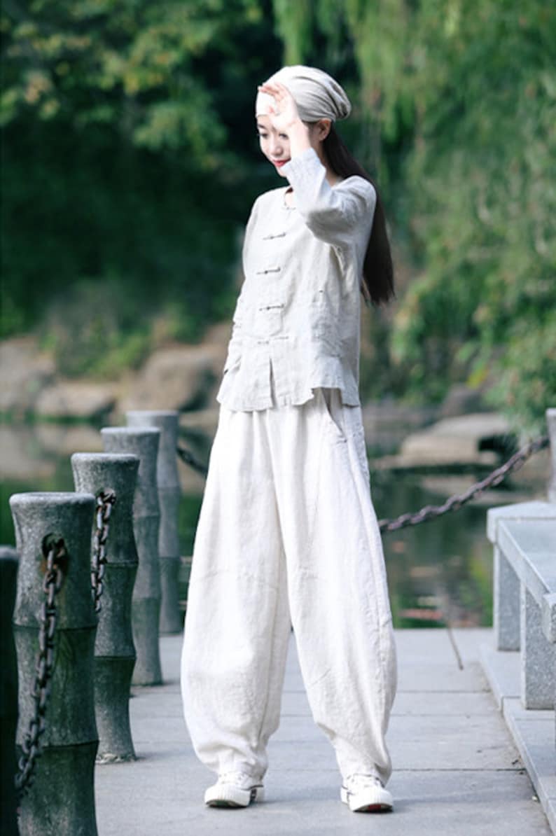 May include: A person wearing a white linen outfit, including a long-sleeved top and wide-leg pants. The outfit is paired with a matching head covering and white sneakers. The person is standing on a stone walkway near a body of water.