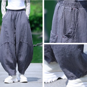 May include: Gray linen pants with a relaxed fit, featuring an elastic waistband and pockets. The pants have a unique, voluminous silhouette and are paired with white sneakers. The fabric appears soft and breathable, suitable for casual wear.