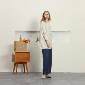 May include: A woman wearing a beige button-down shirt and navy blue pants. The shirt has long sleeves and a relaxed fit. She is standing next to a wooden nightstand with a brown paper bag on top. The bag has text on it.