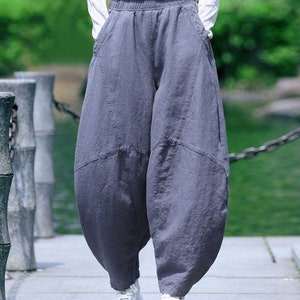 May include: Gray, wide-leg pants with a unique, voluminous shape. The pants have an elastic waistband and pockets. The person is wearing white sneakers and a white long-sleeve shirt. The pants appear to be made of a soft, flowing fabric.