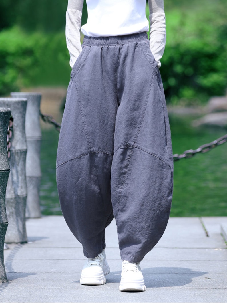 May include: Gray, wide-leg pants with a unique, voluminous shape. The pants have an elastic waistband and pockets. The person is wearing white sneakers and a white long-sleeve shirt. The pants appear to be made of a soft, flowing fabric.