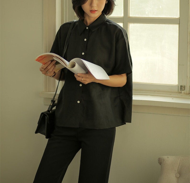 May include: A woman wearing a black collared shirt with short sleeves and black pants. She is holding a magazine in her hands and has a black handbag slung over her shoulder.
