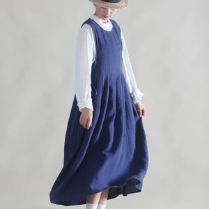 Puede incluir: Una mujer lleva un vestido de lino azul con una blusa blanca debajo. Lleva un sombrero de paja con una cinta negra y calcetines blancos con zapatos marrones.