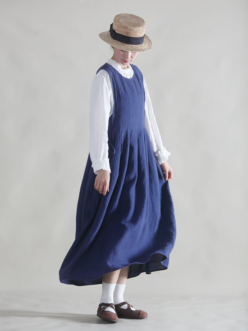 Puede incluir: Una mujer lleva un vestido de lino azul con una blusa blanca debajo. Lleva un sombrero de paja con una cinta negra y calcetines blancos con zapatos marrones.