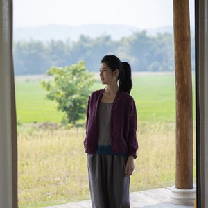May include: A woman wearing a purple cardigan and gray pants stands on a wooden deck overlooking a field of green grass.