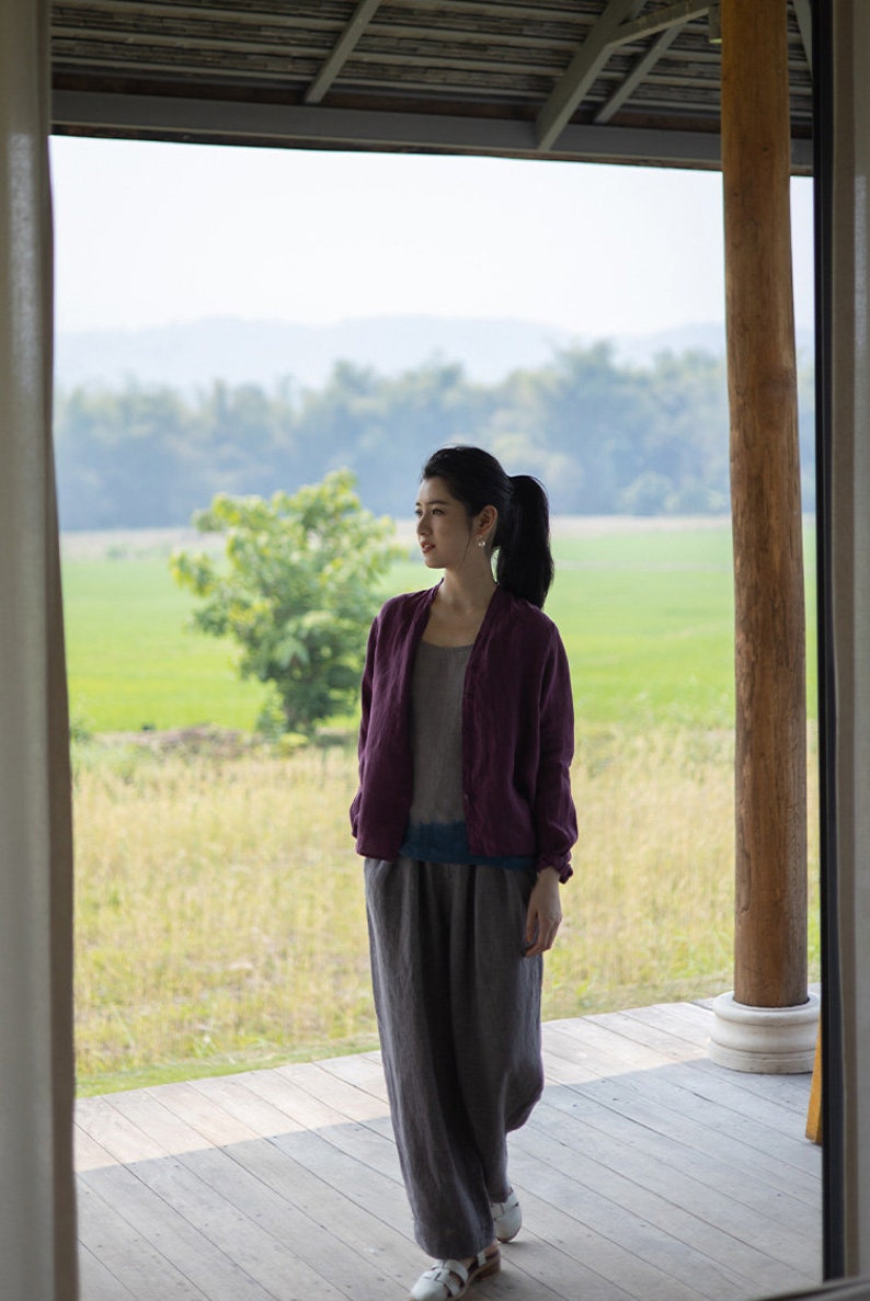 May include: A woman wearing a purple cardigan and gray pants stands on a wooden deck overlooking a field of green grass.
