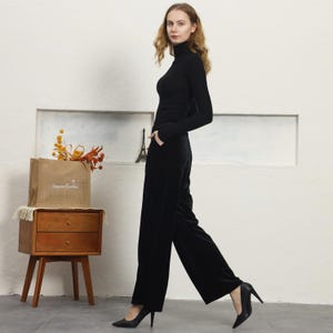 May include: A woman wearing a black turtleneck sweater and black velvet wide-leg pants. She is standing in front of a white wall and is wearing black heels.