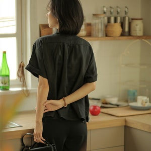 May include: A woman wearing a black linen shirt with short sleeves and black pants. She is holding a black leather handbag with a gold clasp.