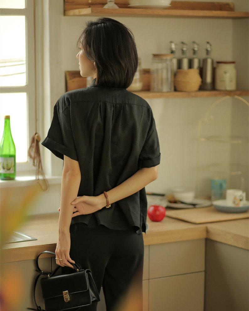 May include: A woman wearing a black linen shirt with short sleeves and black pants. She is holding a black leather handbag with a gold clasp.