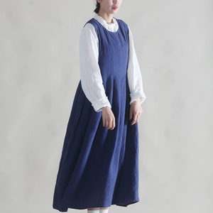Puede incluir: Una mujer con un vestido de lino azul y una blusa blanca debajo. Lleva un sombrero de paja con una cinta negra y zapatos marrones.