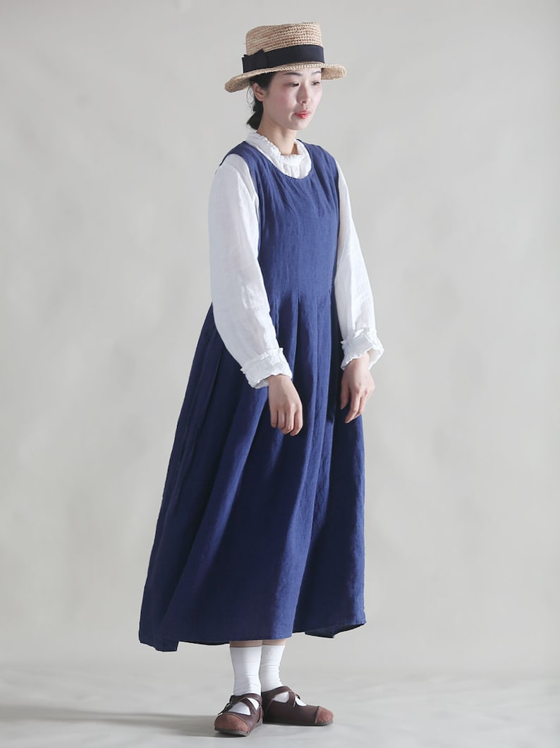 Puede incluir: Una mujer con un vestido de lino azul y una blusa blanca debajo. Lleva un sombrero de paja con una cinta negra y zapatos marrones.