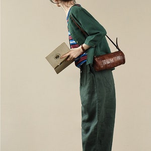 May include: A woman wearing a green linen shirt and pants with a brown leather crossbody bag. She is holding a book and wearing white sneakers.